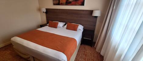 A hotel room with a large bed, two bedside tables, a headboard, a painting on the wall, and a window with curtains.