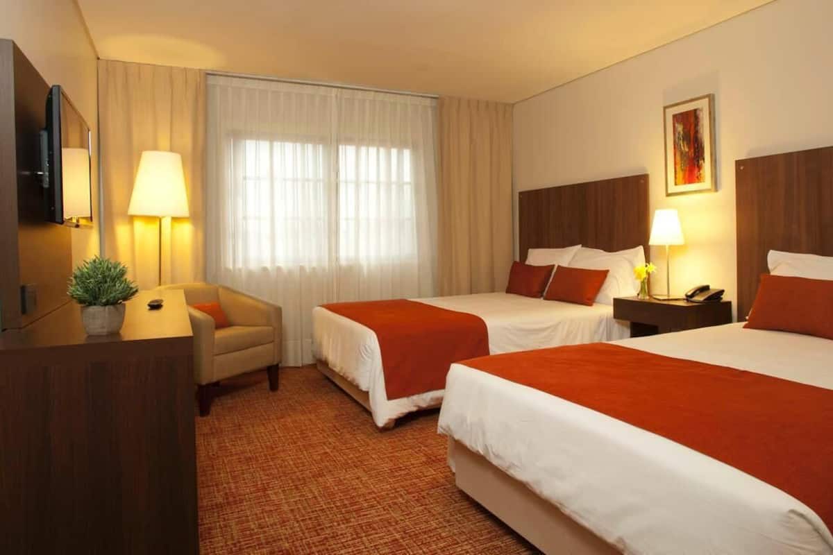 A hotel room with two beds, a desk, a chair, a TV, and a window with curtains.