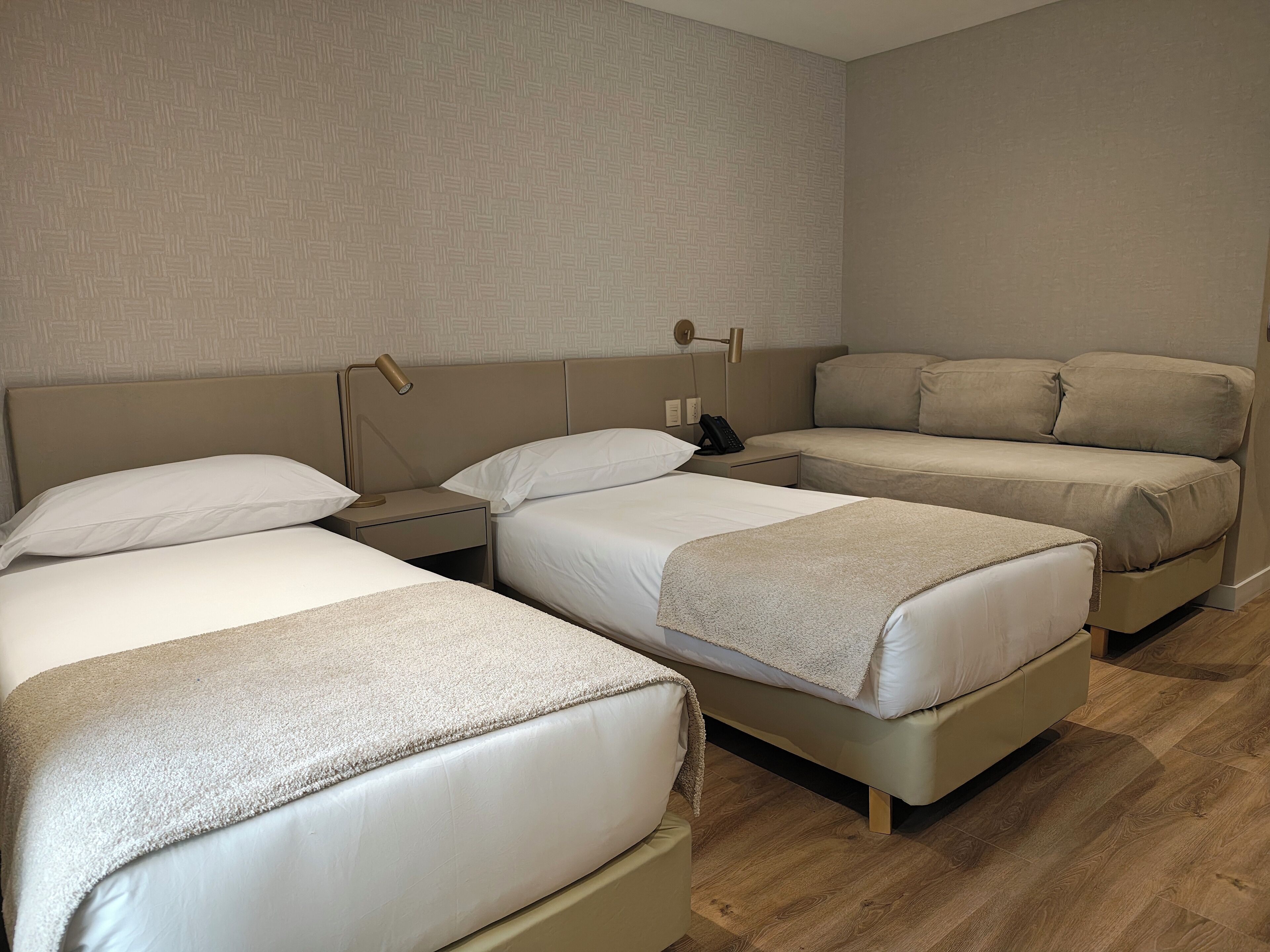 Habitación de hotel con tres camas, una mesita de noche con lámpara y un teléfono en la pared.