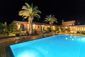 A swimming pool with lounge chairs and palm trees, surrounded by a building with outdoor seating and a stone pathway. - Hotel Masseria Le Pajare (Presicce-Acquarica)