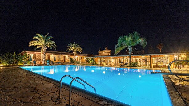 A swimming pool with a stone deck, palm trees, and a building with a warm exterior lighting. - Hotel Masseria Le Pajare (Presicce-Acquarica)