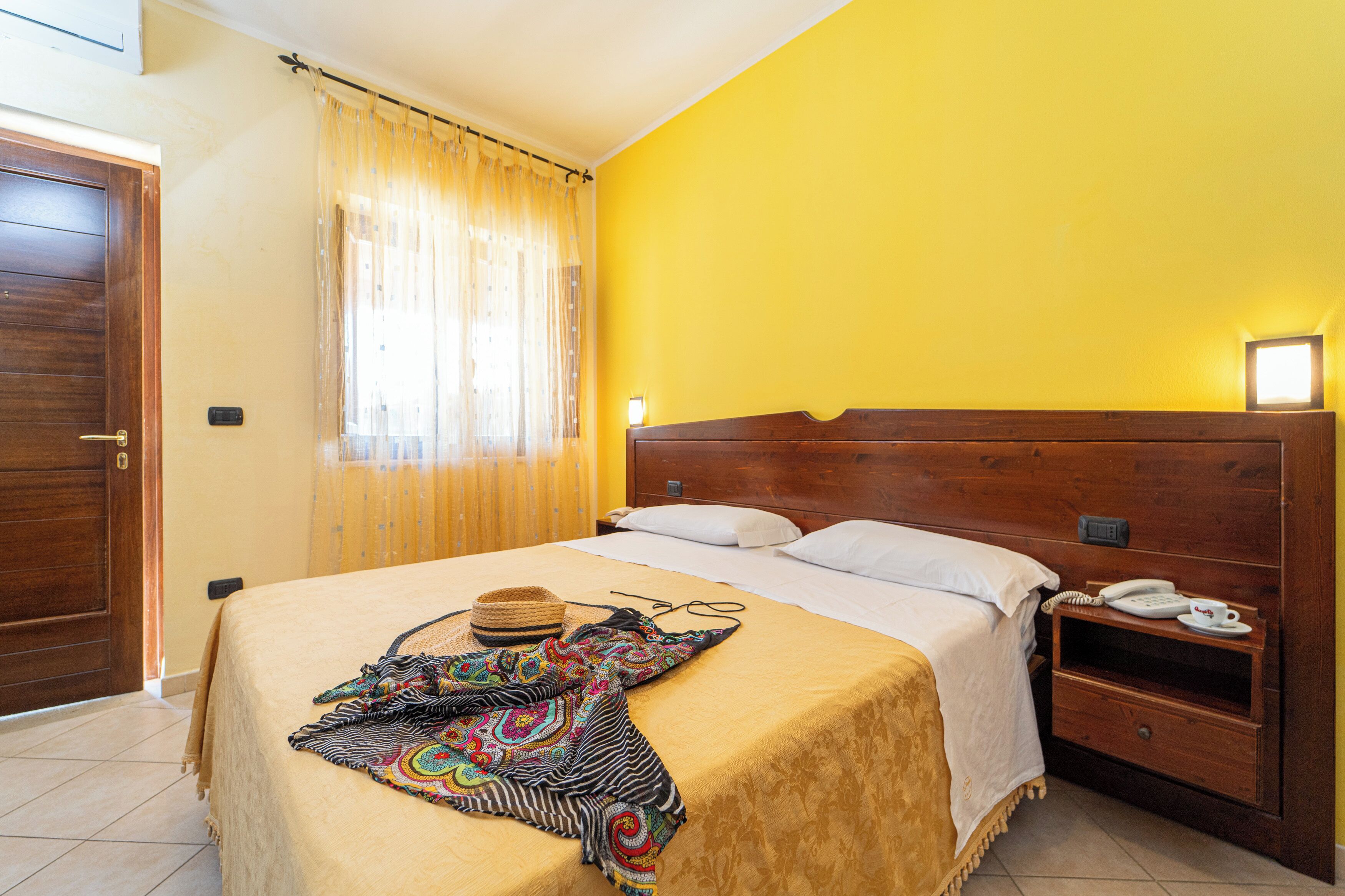 Ein Hotelzimmer mit einem hölzernen Kopfteil, einem Bett mit beiger Bettdecke, einer bunten Decke, einem Hut und einem Nachttisch mit Telefon.