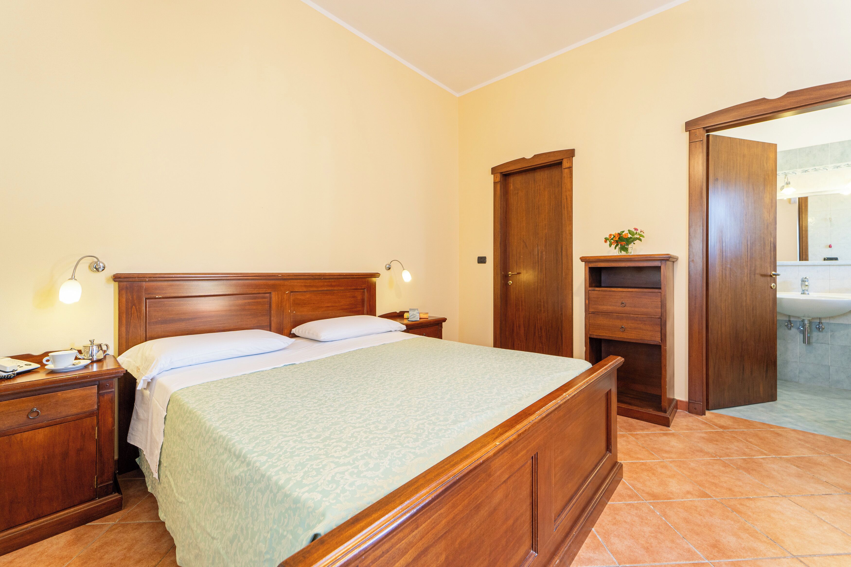 Habitación de hotel con cama de madera, mesitas de noche, cómoda y baño con lavamanos y espejo.