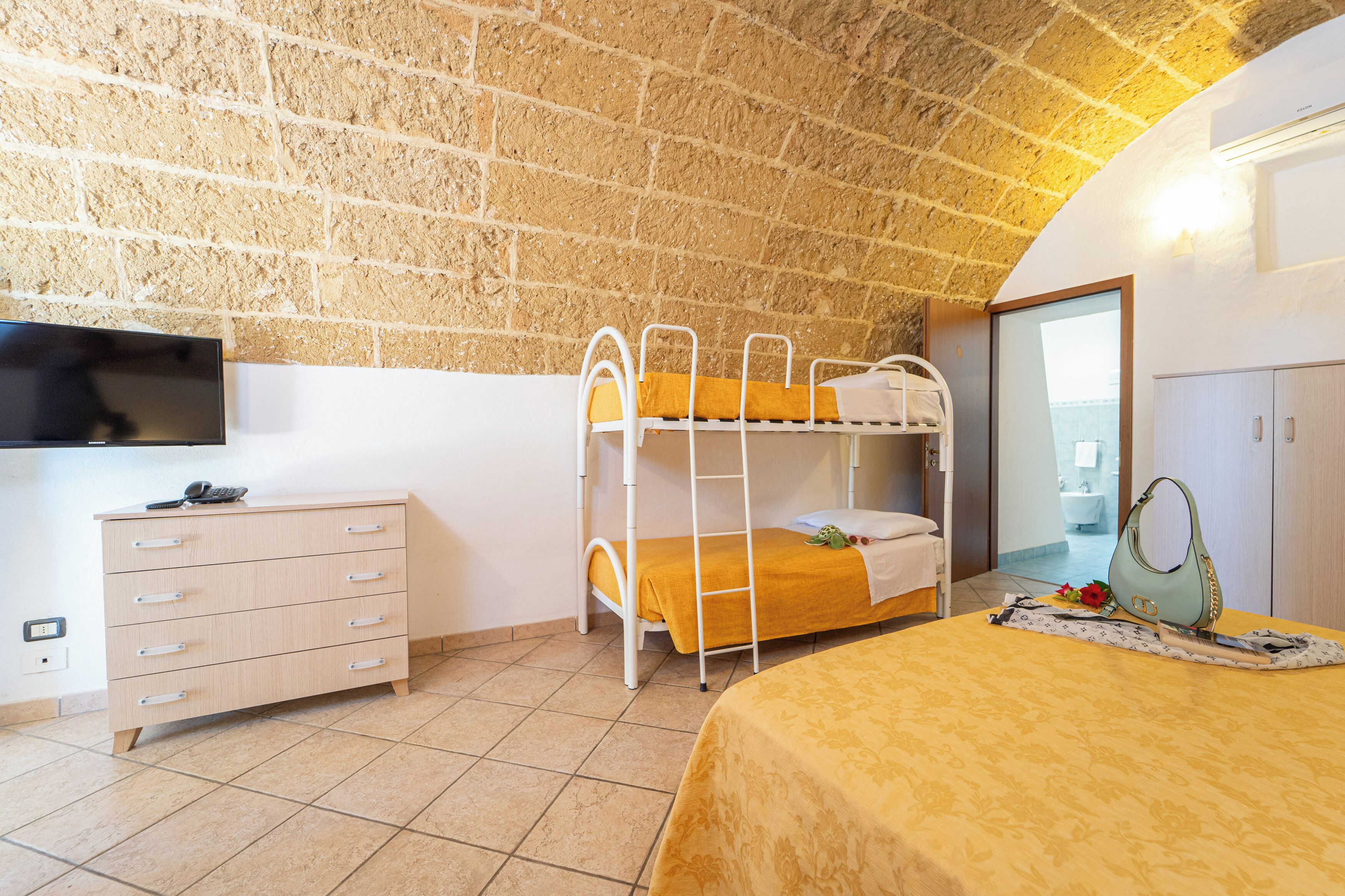 Una stanza con un letto a castello, una cassettiera, una televisione e l'ingresso del bagno.