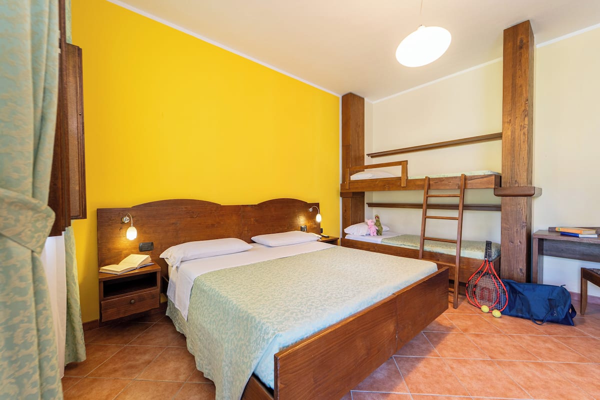A bedroom with a bunk bed, a bedside table with a lamp, a desk with a chair, and a tennis racket bag.