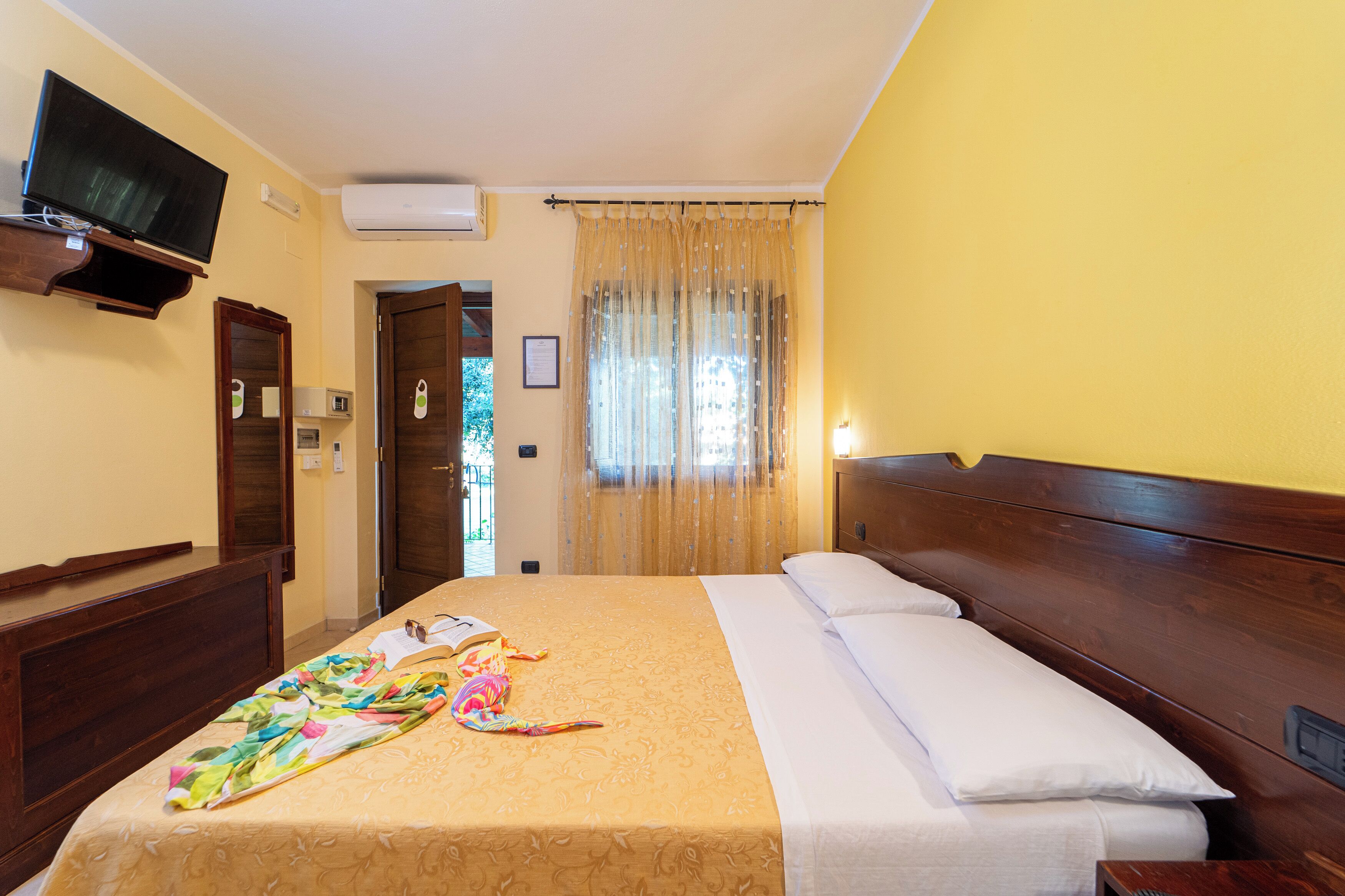 Habitació d'hotel amb llit, televisió penjada a la paret, panell de fusta al capçal i finestra amb cortines.