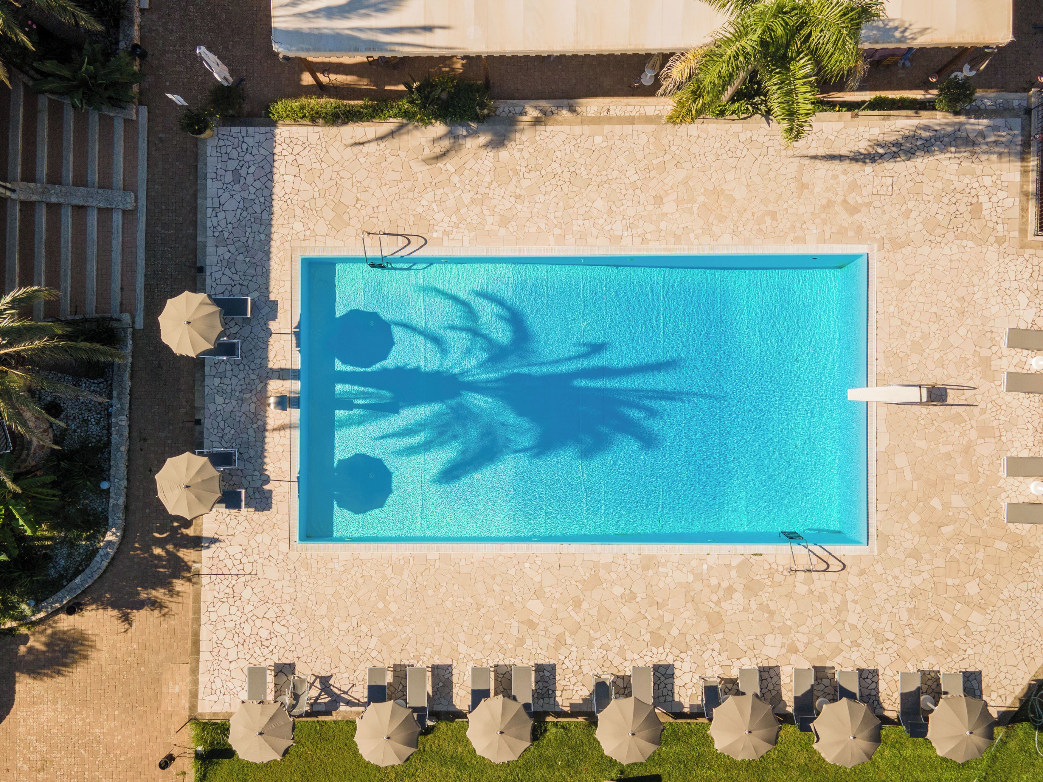 An aerial view of a rectangular swimming pool with a tiled deck surrounding it, equipped with multiple lounge chairs and umbrellas.