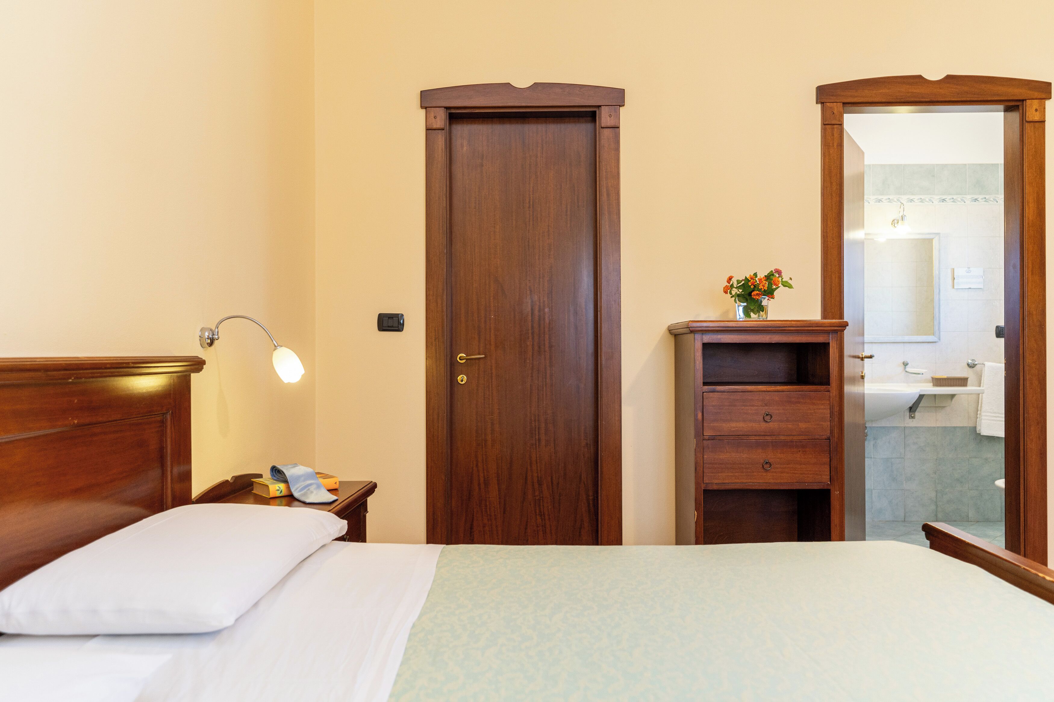 A neatly arranged hotel room with a wooden bed, a nightstand with a lamp, a wooden wardrobe, and a bathroom visible through an open door.