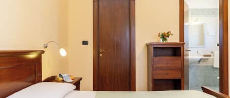 A neatly arranged hotel room with a wooden bed, a nightstand with a lamp, a wooden wardrobe, and a bathroom visible through an open door.