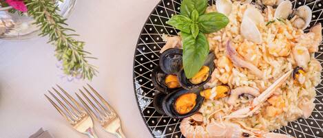Un plato de risotto de mariscos con mejillones, almejas y camarones, adornado con albahaca, sobre un plato negro con estampado de lunares blancos.