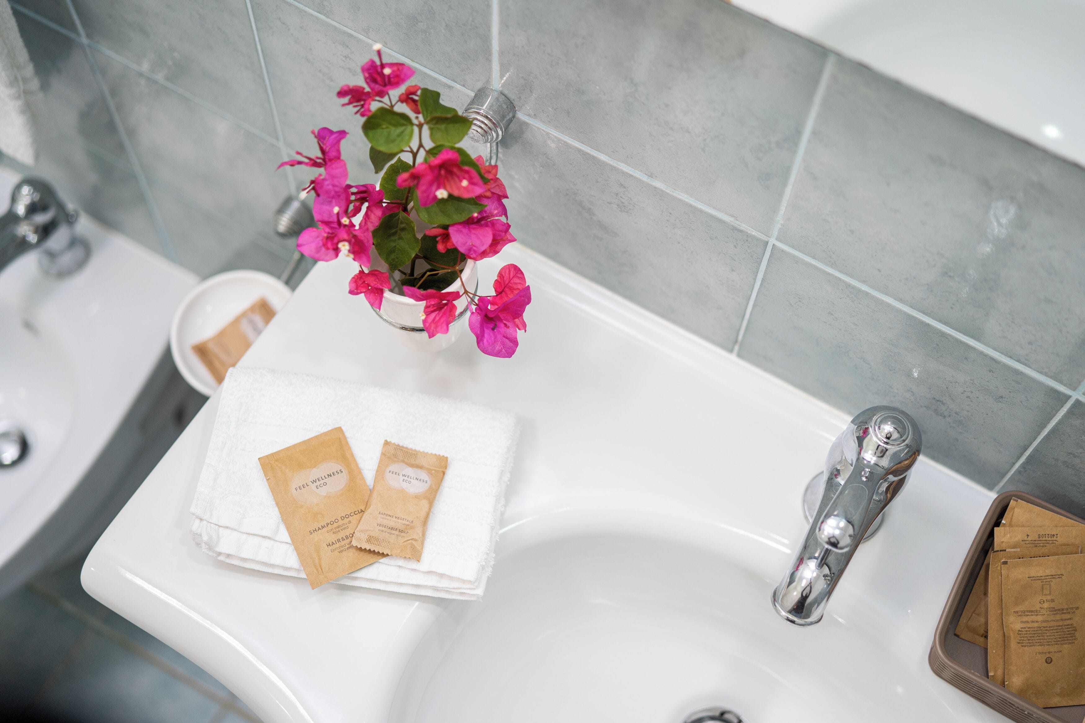 A bathroom sink with a white towel, two hair care packets, and a potted plant with pink flowers.