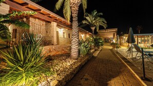 A nighttime view of a hotel corridor with stone walls, palm trees, and outdoor seating. - Hotel Masseria Le Pajare (Presicce-Acquarica)