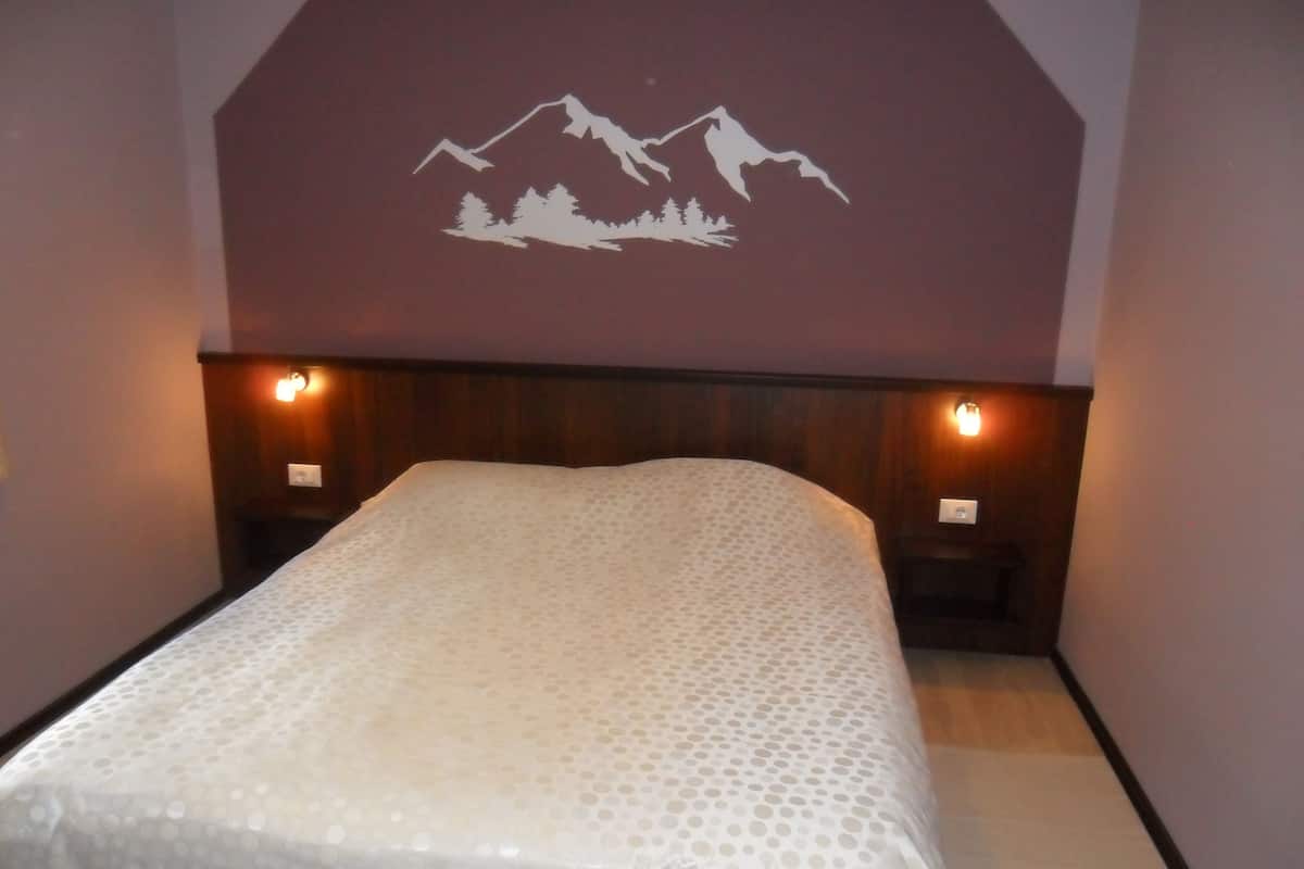 A bedroom with a bed, wooden headboard, two bedside lamps, and a wall mural of mountains.