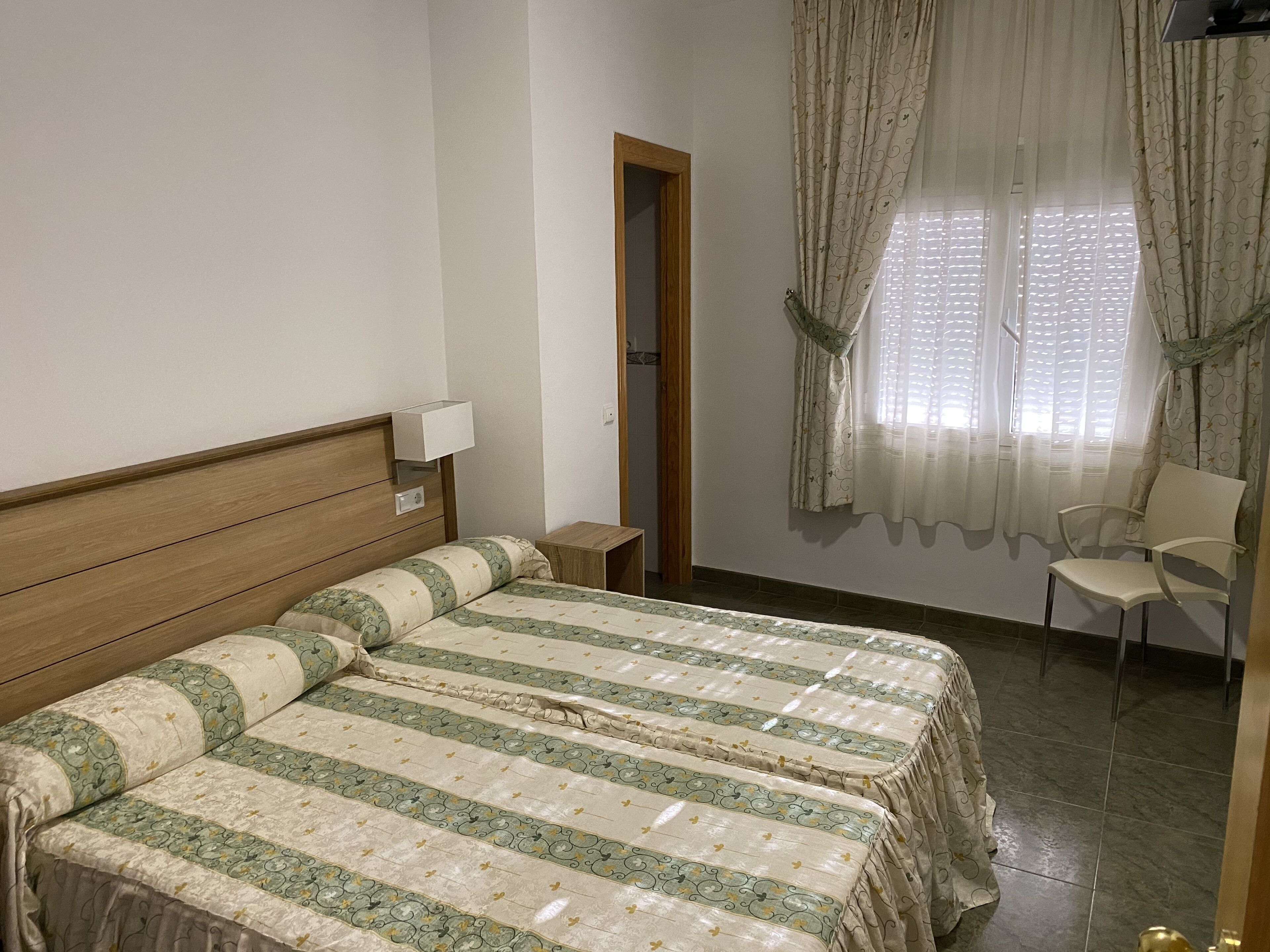 Un dormitorio con cama, mesita de noche, silla y ventana con cortinas.