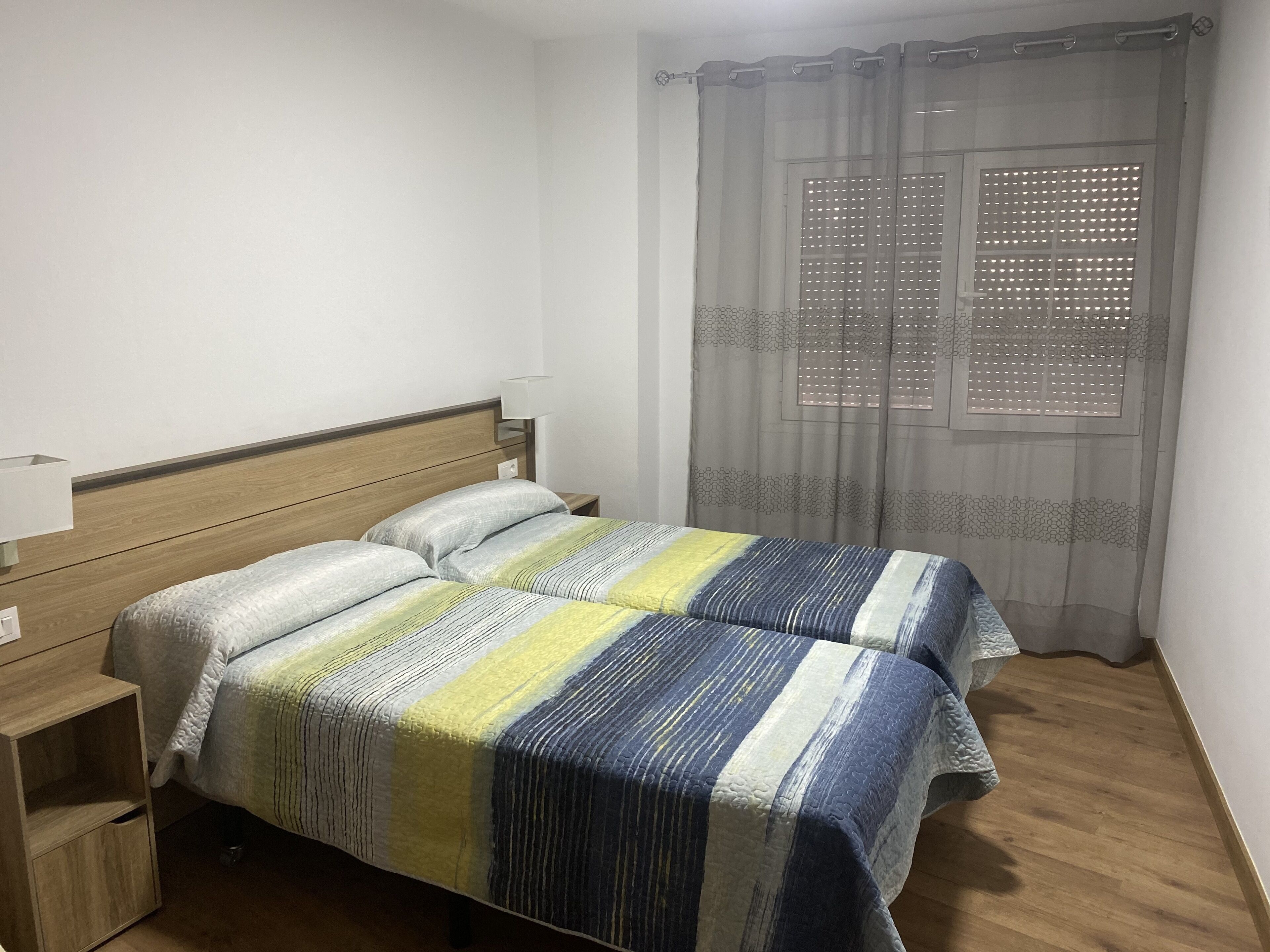 A bedroom with two beds, a wooden headboard, a bedside table with a lamp, and a window with a patterned curtain.