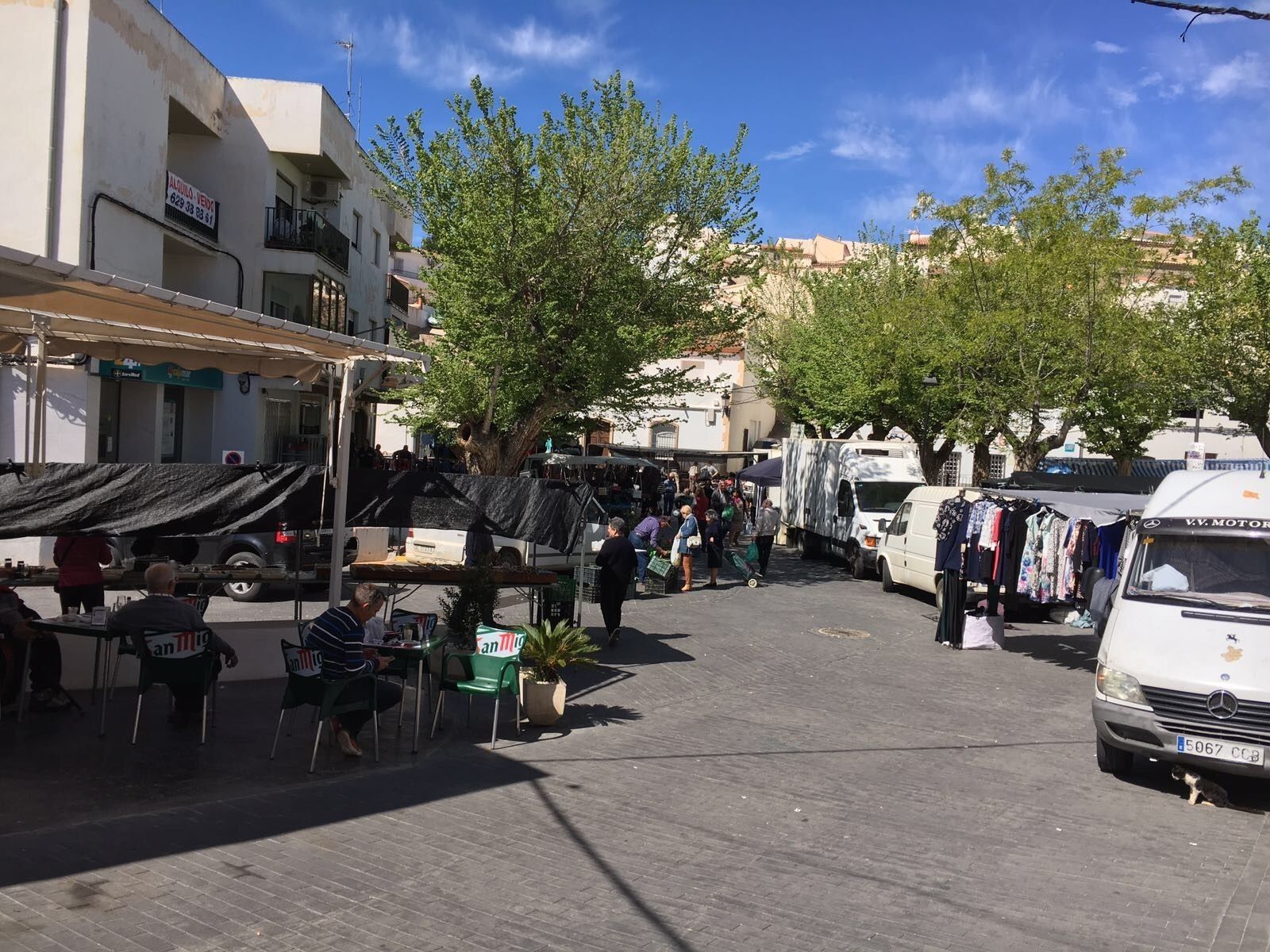 Un mercado callejero con vendedores y compradores, una furgoneta estacionada y un árbol en el centro.