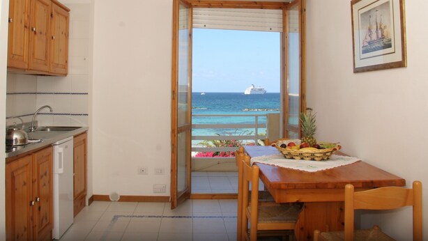 A kitchen with wooden cabinets, a white refrigerator, and a dining table with chairs. A window opens to a balcony with a view of the sea and a ship.