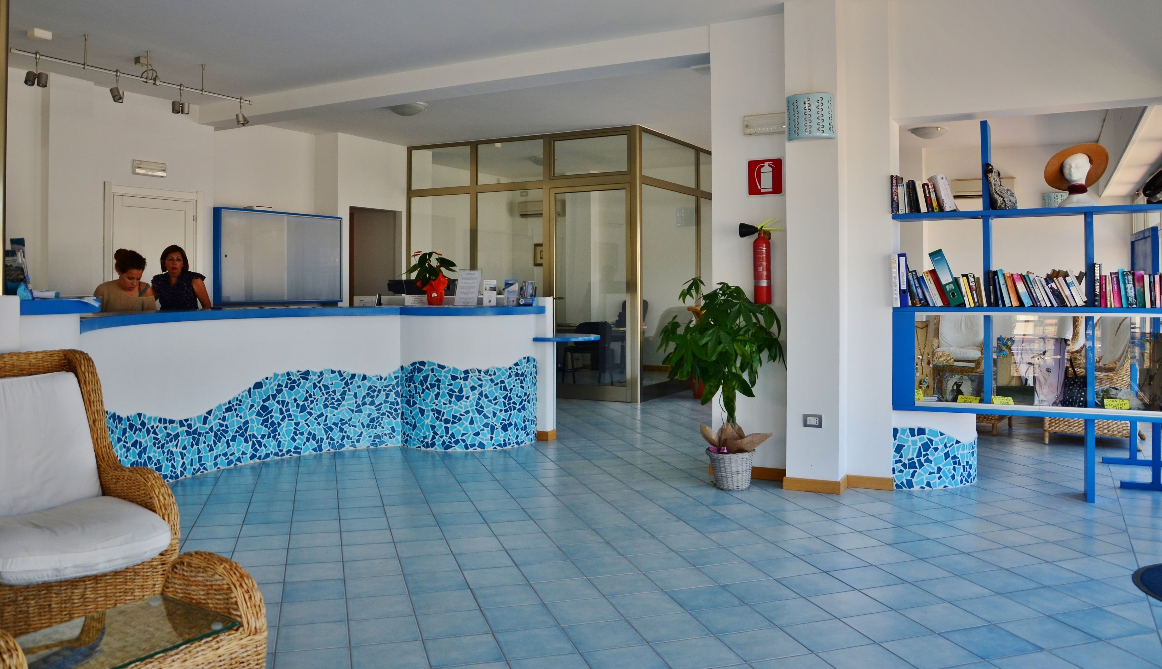 A reception area with a blue tiled floor, a reception desk, a wicker chair, a bookshelf, and a potted plant.