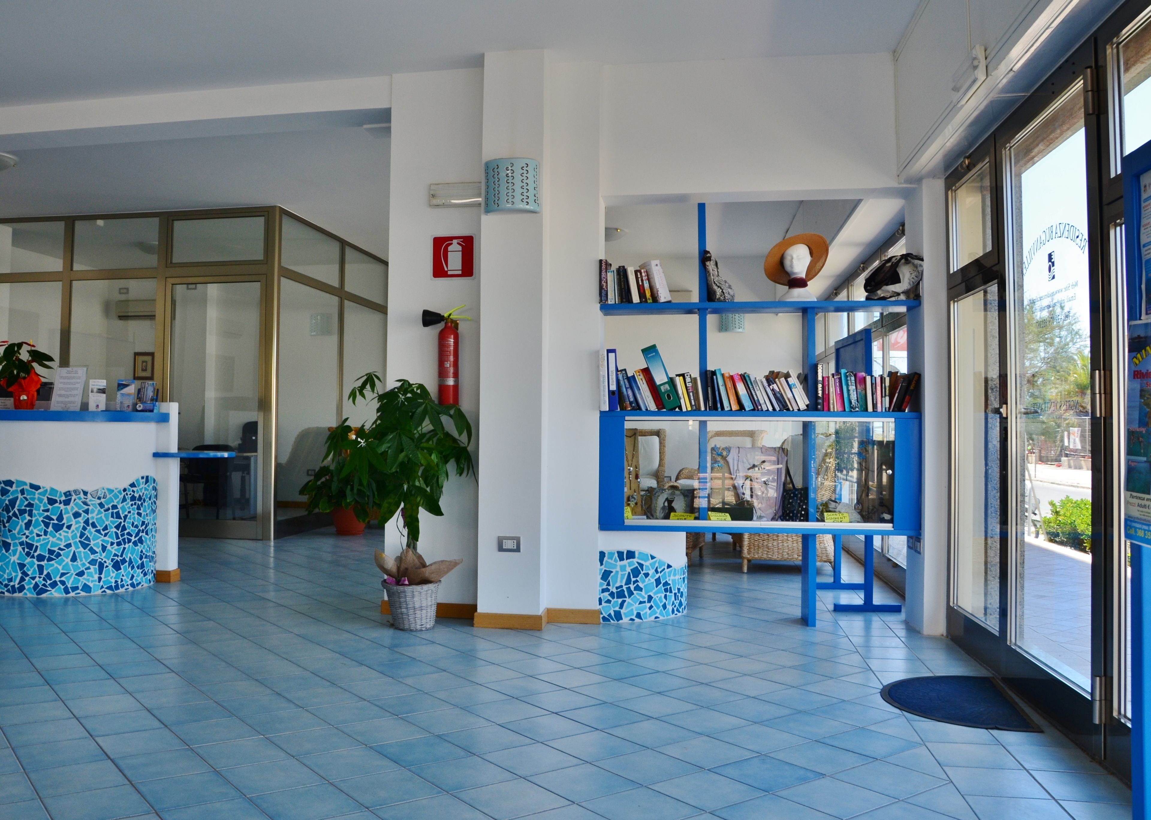 Aula, jossa on sinisillä laatoilla päällystetty lattia, vastaanottotiski, ruukussa oleva kasvi, kirjahylly ja palosammutin.