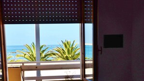A room with a bed, a window with a view of the ocean and palm trees, and a small table with a towel.