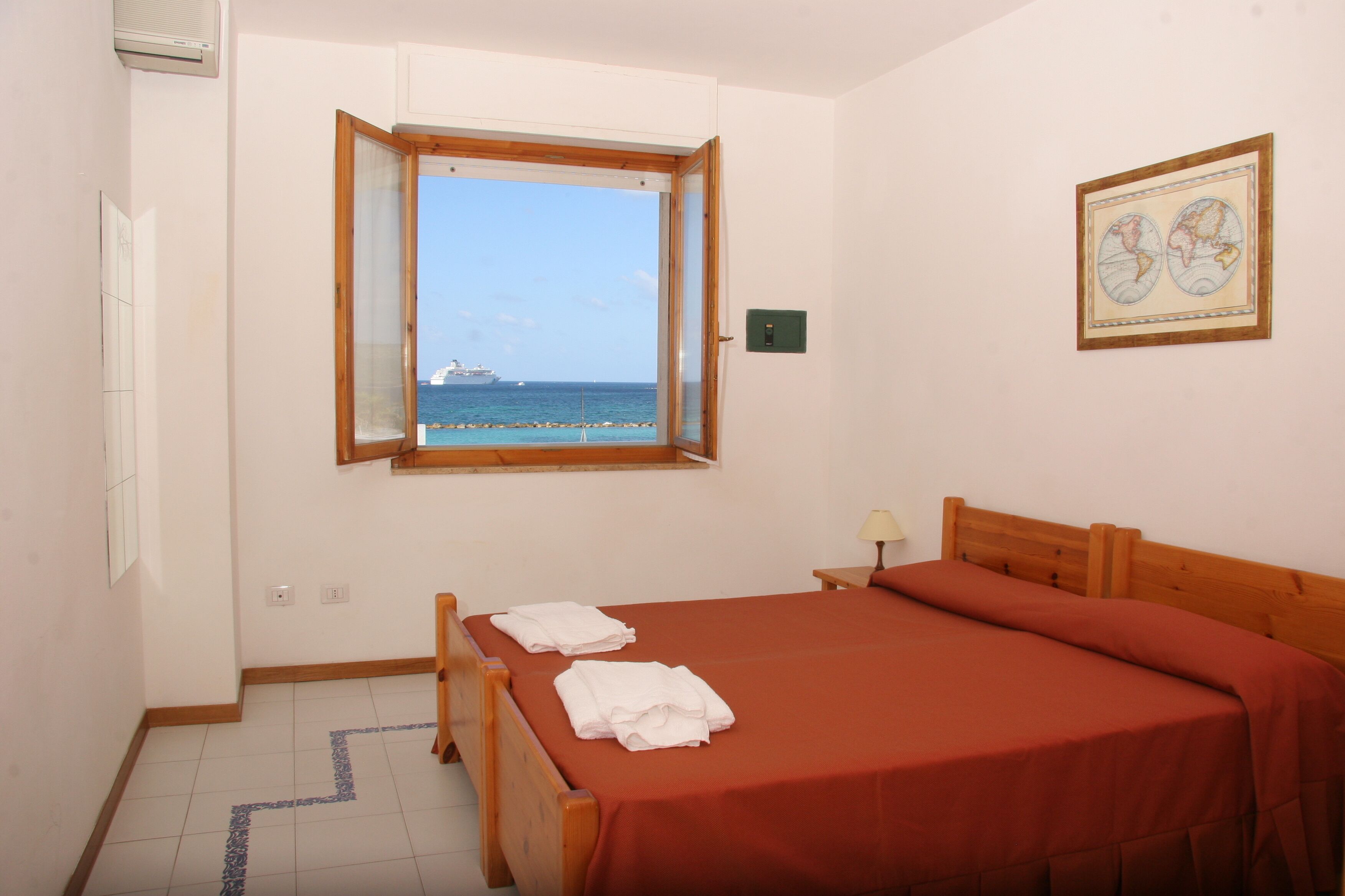 A bedroom with a large bed, a window with a view of the sea and a ship, and a framed map on the wall.