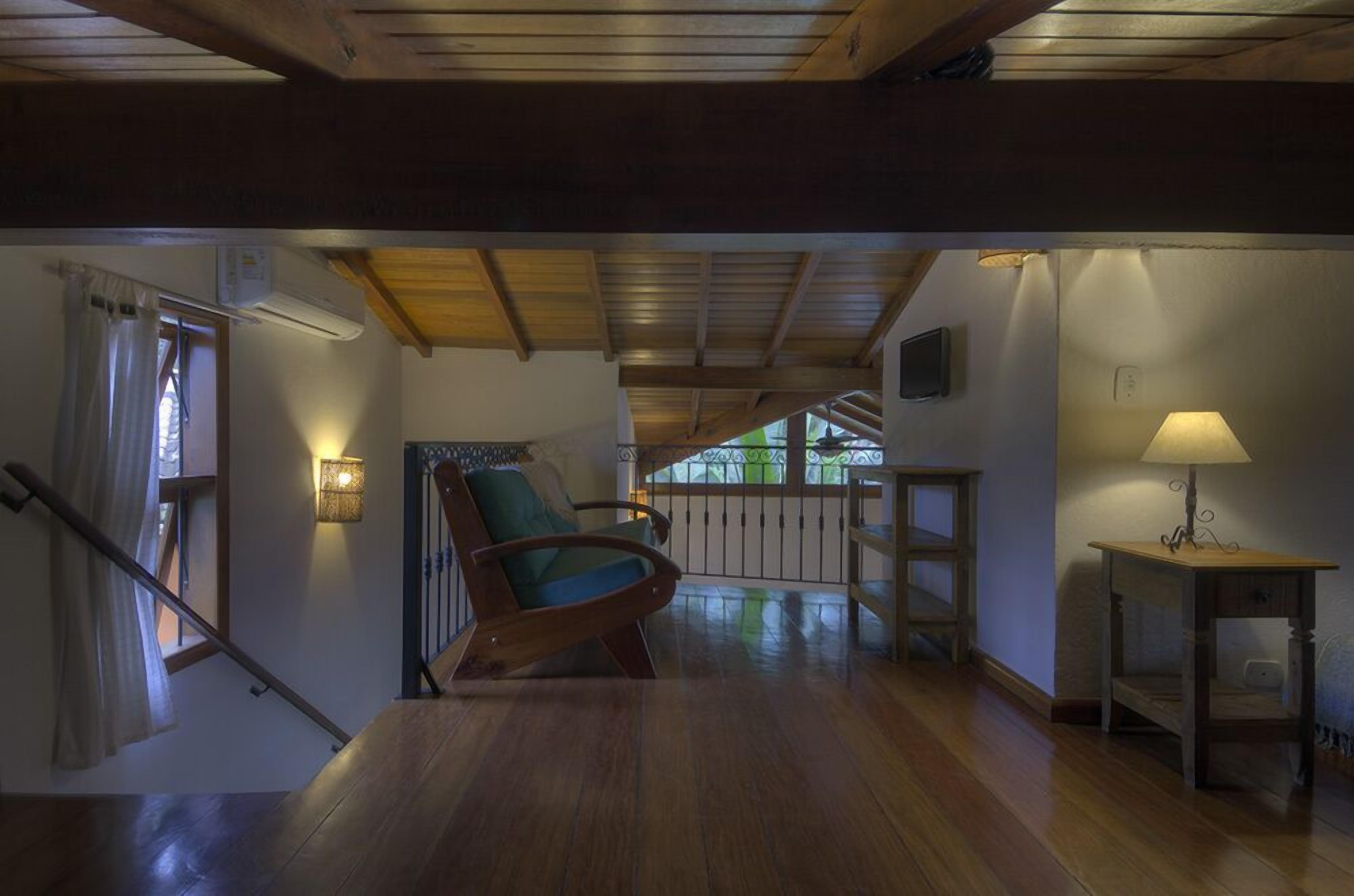 Amplia habitación con suelo de madera, una silla de madera, una mesa con lámpara y una escalera que lleva a otro nivel.