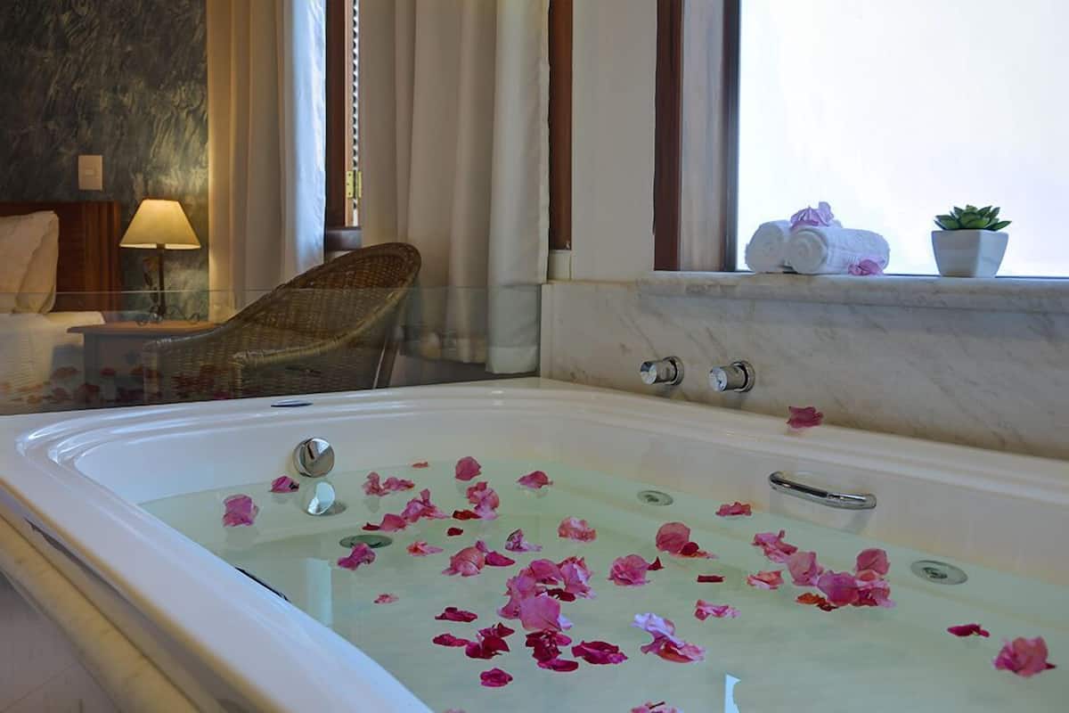 A bathroom with a large bathtub filled with rose petals, a wicker chair, and a small potted plant on a windowsill.