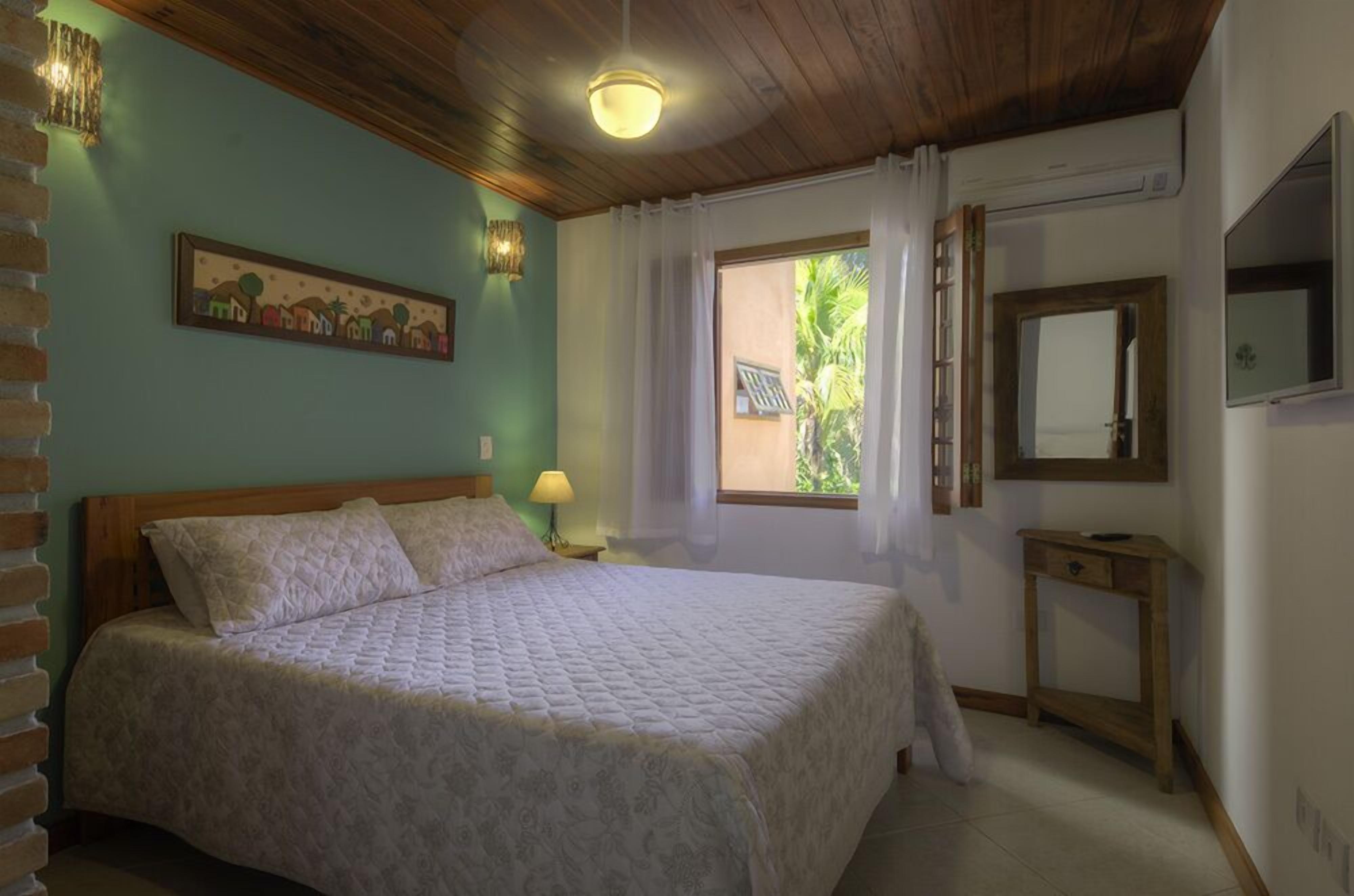 A bedroom with a bed, a bedside lamp, a window with curtains, a wooden headboard, and a wall-mounted mirror.