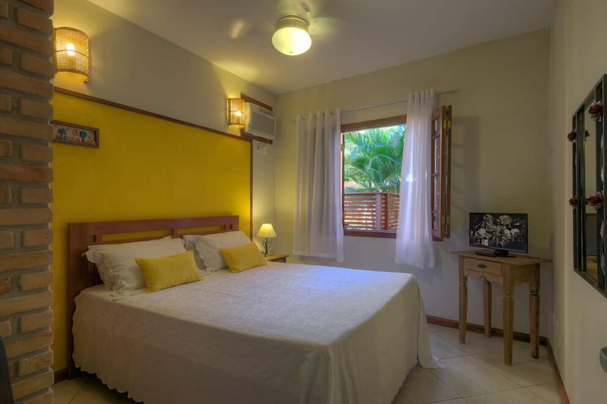 A bedroom with a bed, a nightstand, a lamp, a window with curtains, and a small table with a picture frame.