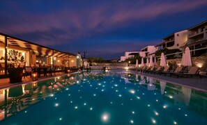 A swimming pool with lounge chairs and umbrellas, surrounded by buildings with outdoor seating and a sunset sky.