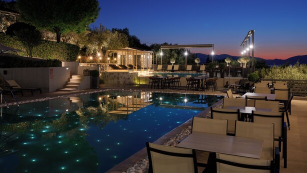 A nighttime pool area with seating and a covered lounge, surrounded by trees and a mountain view. - Altamar Hotel (Istiaia-Aidipsos)