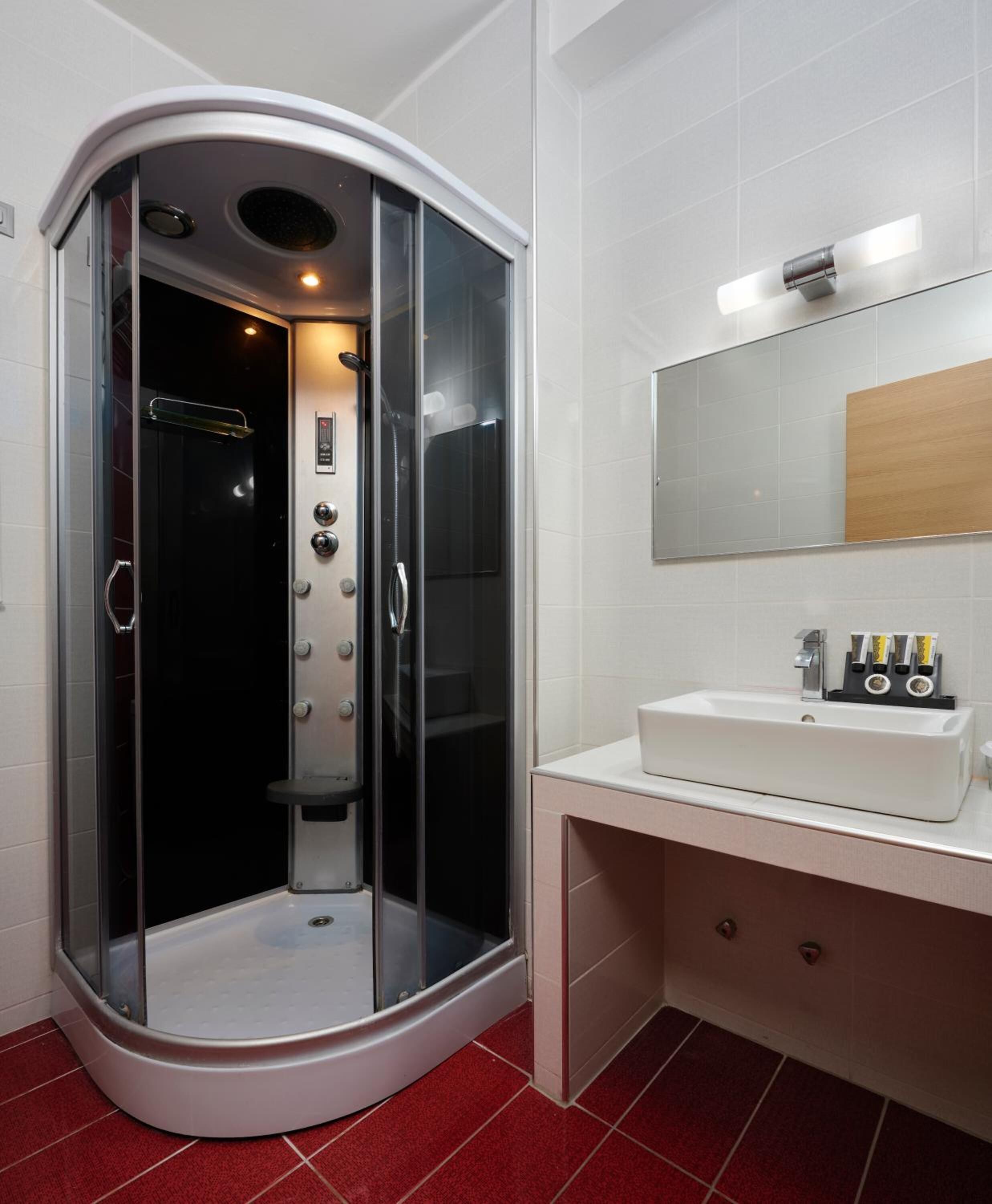 Une salle de bain moderne avec une douche à l’italienne en verre, un lavabo blanc avec un miroir au-dessus, et un sol carrelé de carreaux rouges.