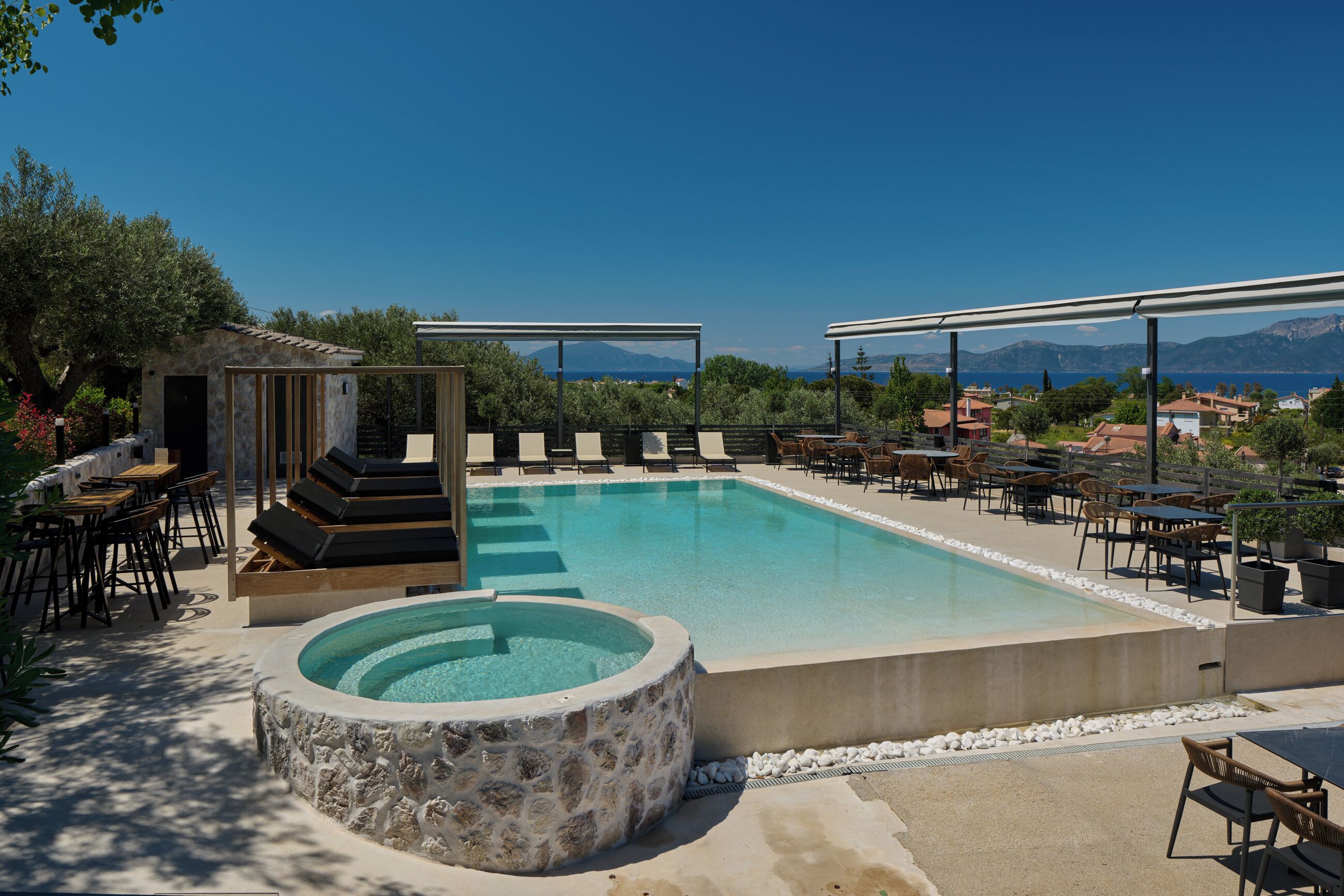 Un área de piscina con sillones reclinables y un jacuzzi, rodeada de mesas y sillas, con vistas al mar y las montañas.