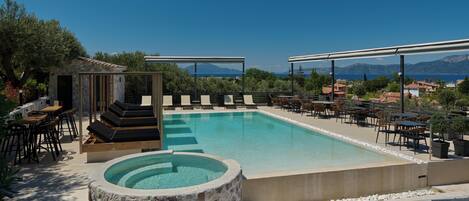 Un área de piscina con sillones reclinables y un jacuzzi, rodeada de mesas y sillas, con vistas al mar y las montañas.