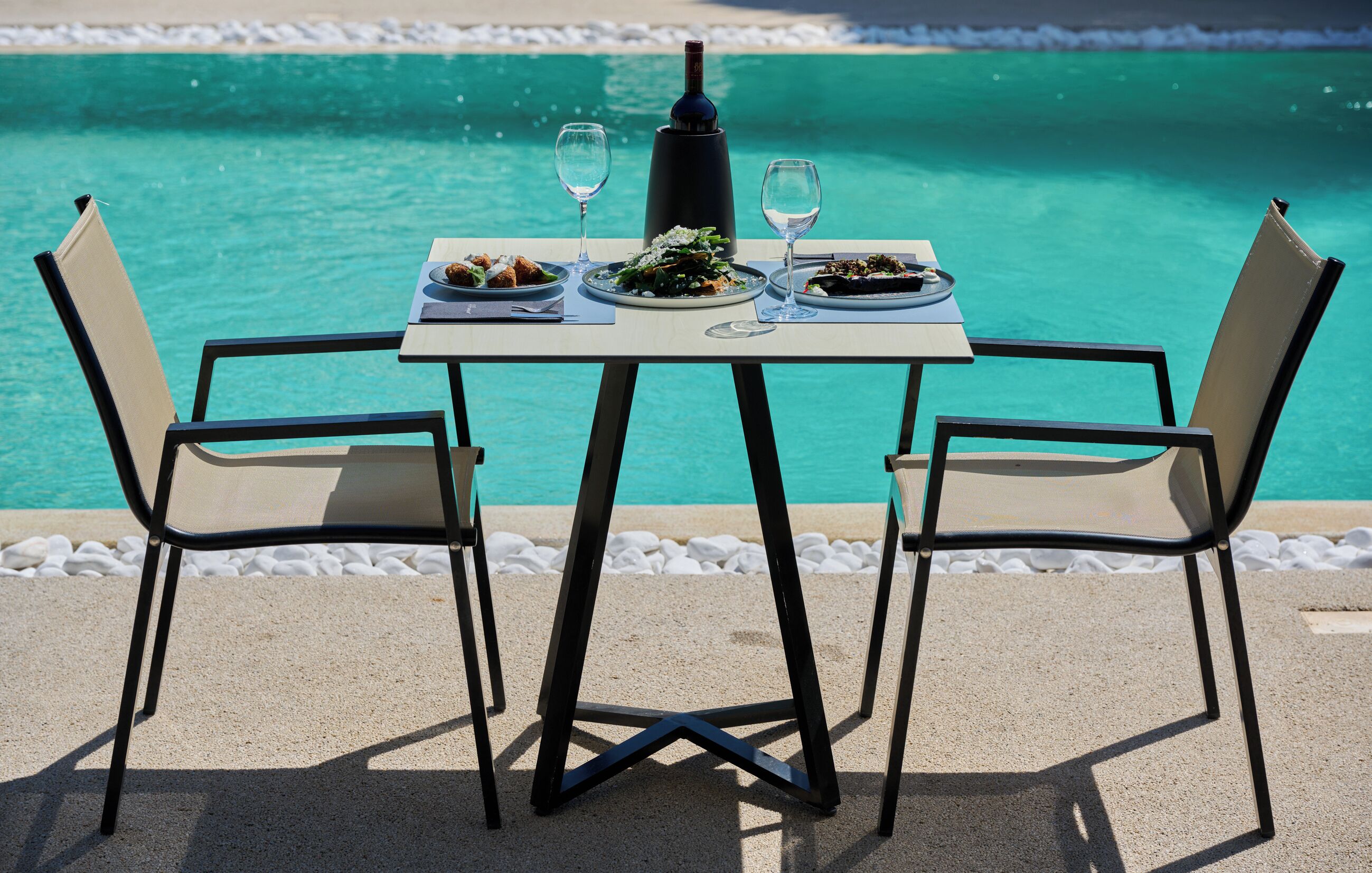 Mesa para dos con platos de comida y copas de vino, con vista a un océano turquesa.