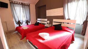 Habitación de hotel con dos camas, cada una con una colcha roja y un cojín blanco. Hay una mesita de noche con una lámpara y una pequeña mesa con una toalla doblada. El piso es de madera y hay un radiador en la pared.