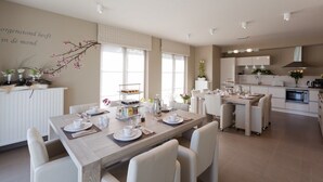 A modern kitchen with white cabinetry, a central island, and a dining area with a wooden table and white chairs. - B&B Riche Terre (Damme)