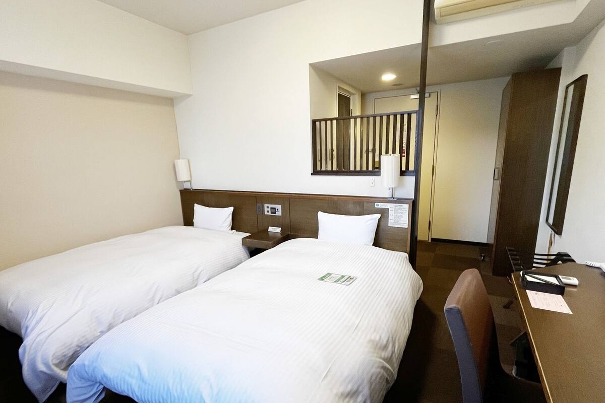 A hotel room with two beds, a desk, a chair, a mirror, and a small kitchenette.