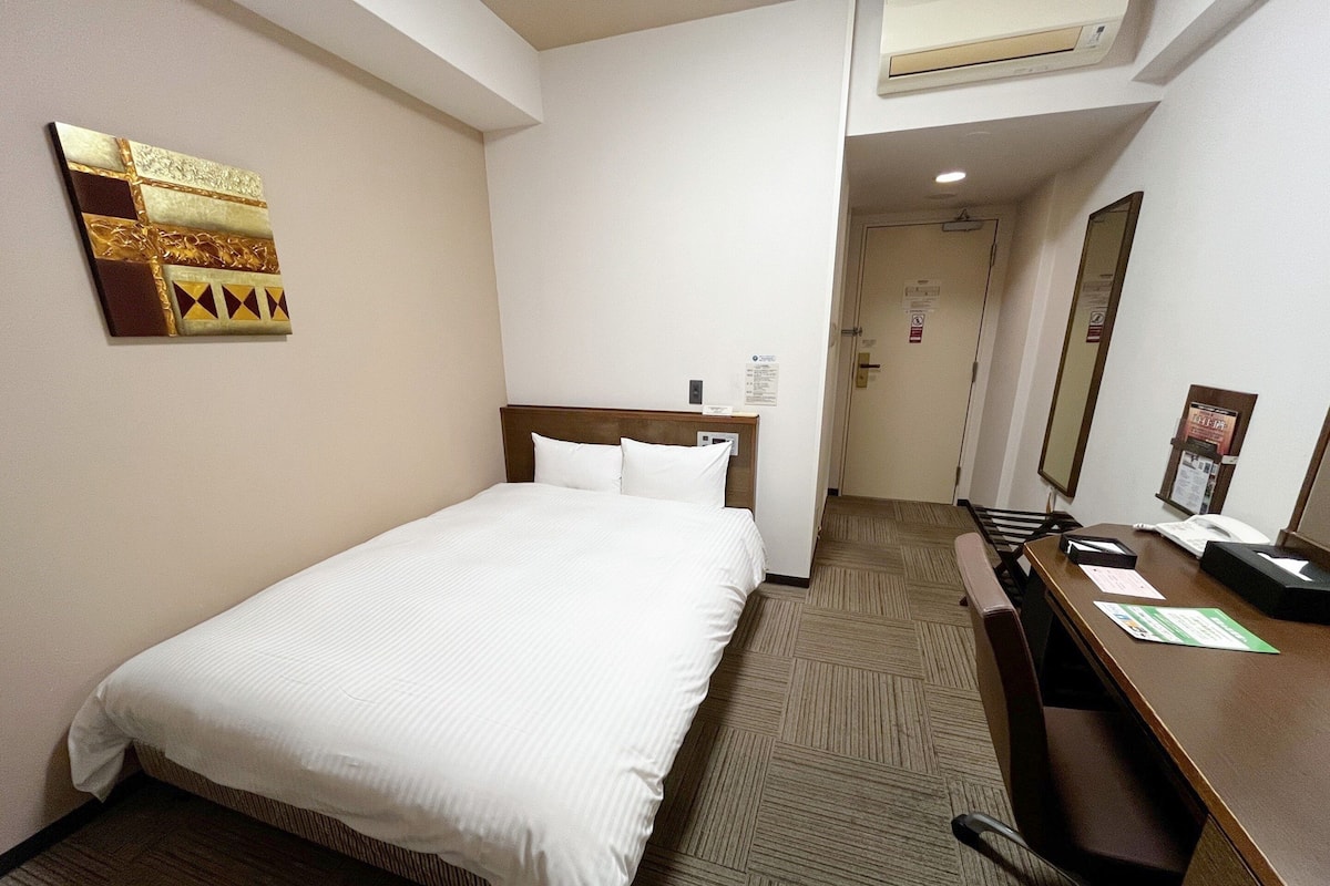 A hotel room with a bed, a desk, a chair, a mirror, and a wall-mounted artwork.