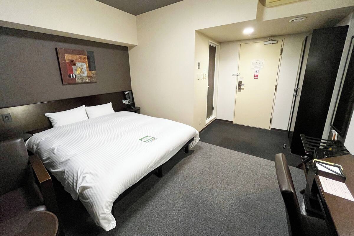 A hotel room with a bed, a desk, a chair, a nightstand, a wall-mounted TV, and a door.