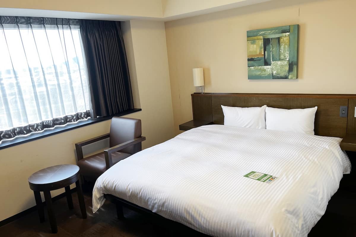 A hotel room with a large bed, a desk, a chair, a window with curtains, a painting on the wall, and a nightstand with a lamp.
