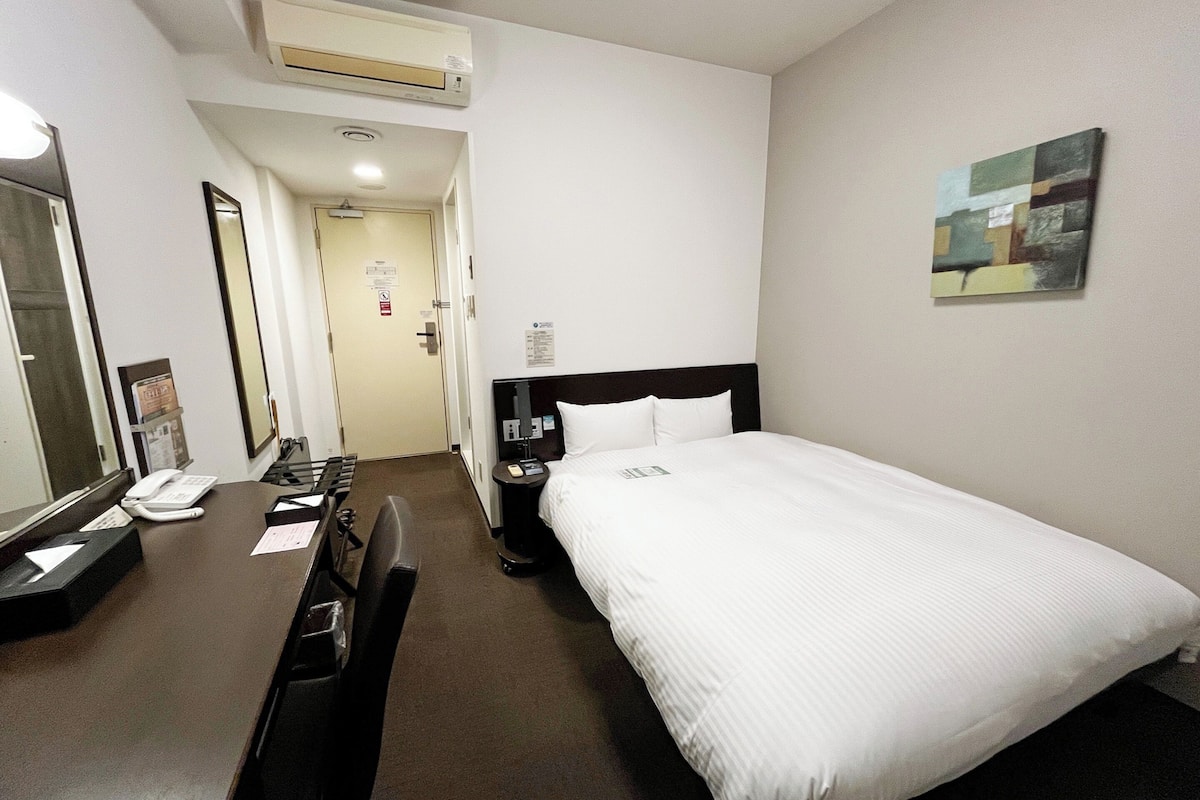 A hotel room with a bed, a desk with a telephone, a chair, a bedside table with a lamp, and a painting on the wall.