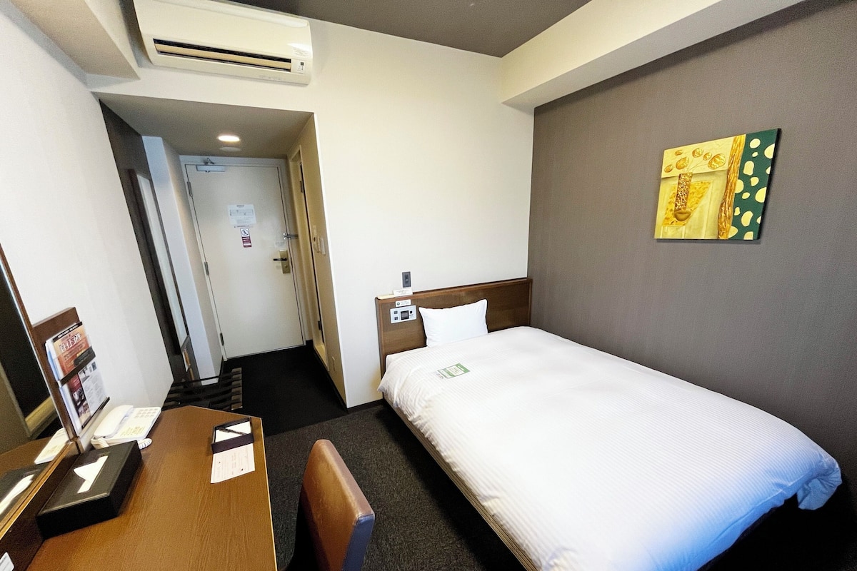A hotel room with a single bed, a desk with a chair, a wooden headboard, a wall-mounted air conditioner, and a door leading to another room.