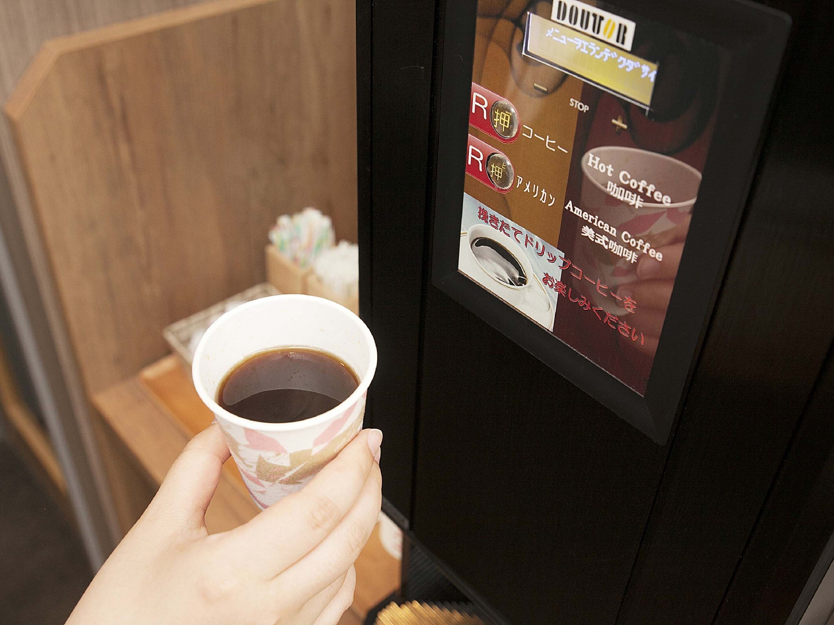 A hand holding a cup of coffee with a digital menu display on a coffee machine.