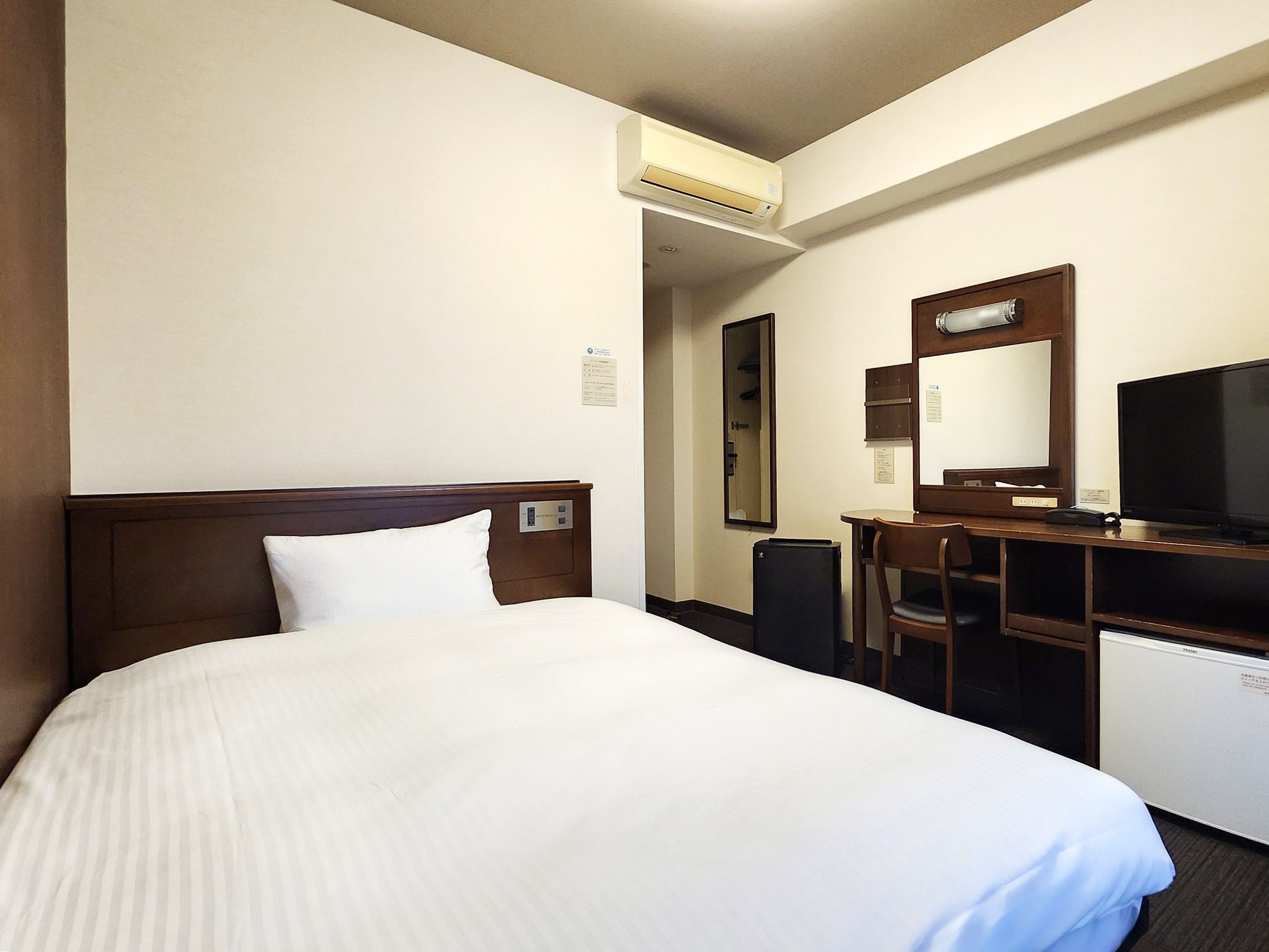 A hotel room with a bed, a desk with a chair, a television, and an air conditioning unit.