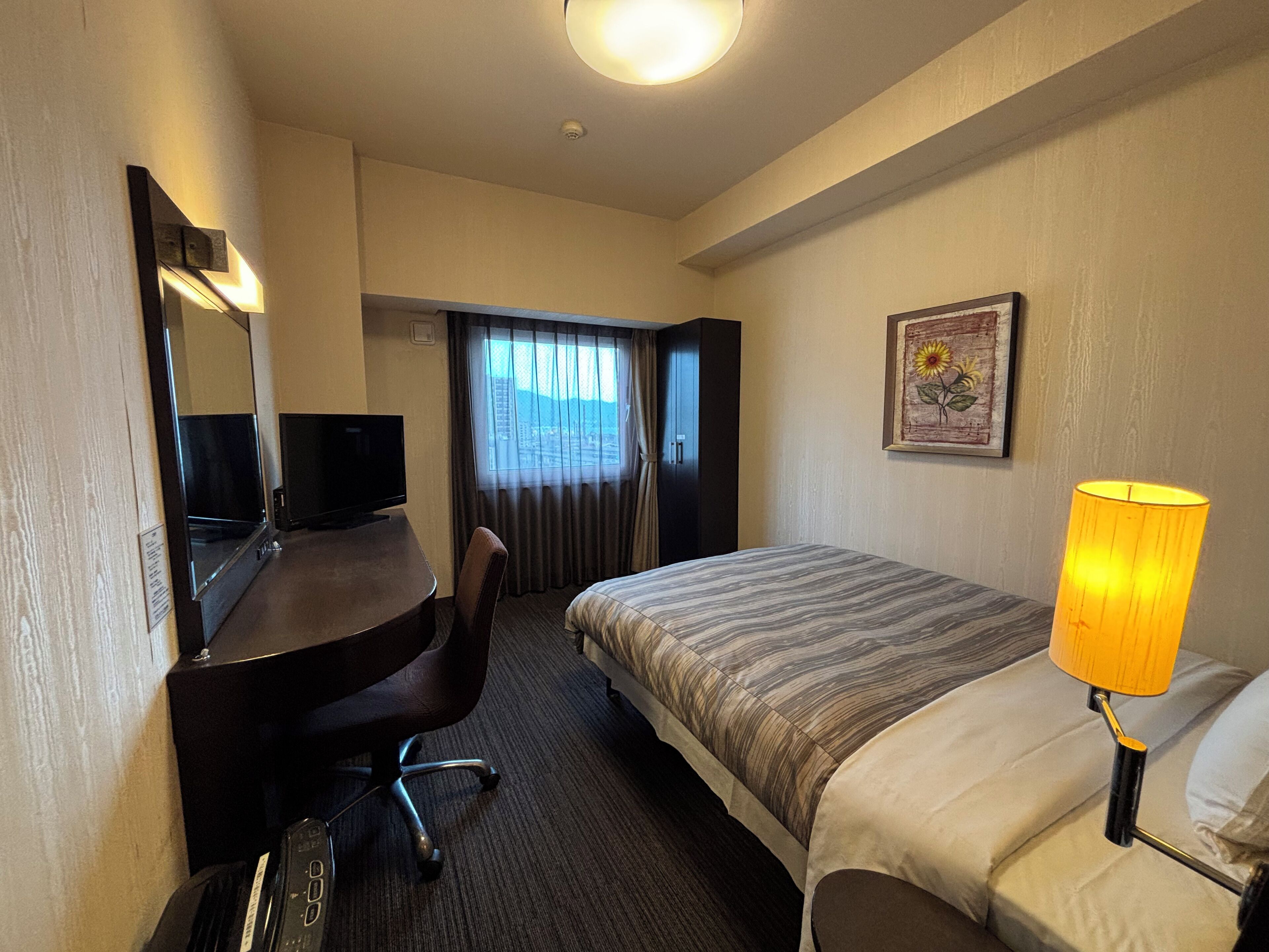 A hotel room with a bed, a desk with a chair, a television, a lamp, and a window with curtains.