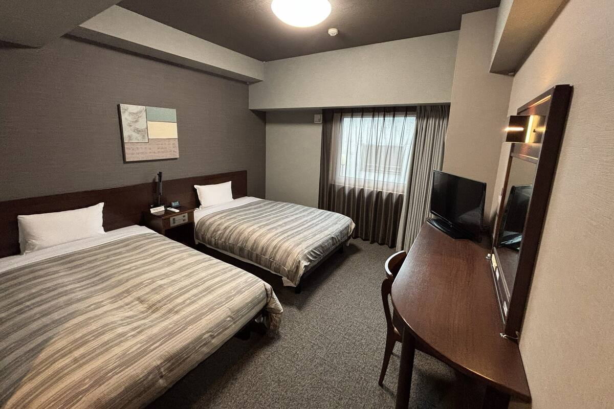 A hotel room with two beds, a desk, a television, and a window with curtains.