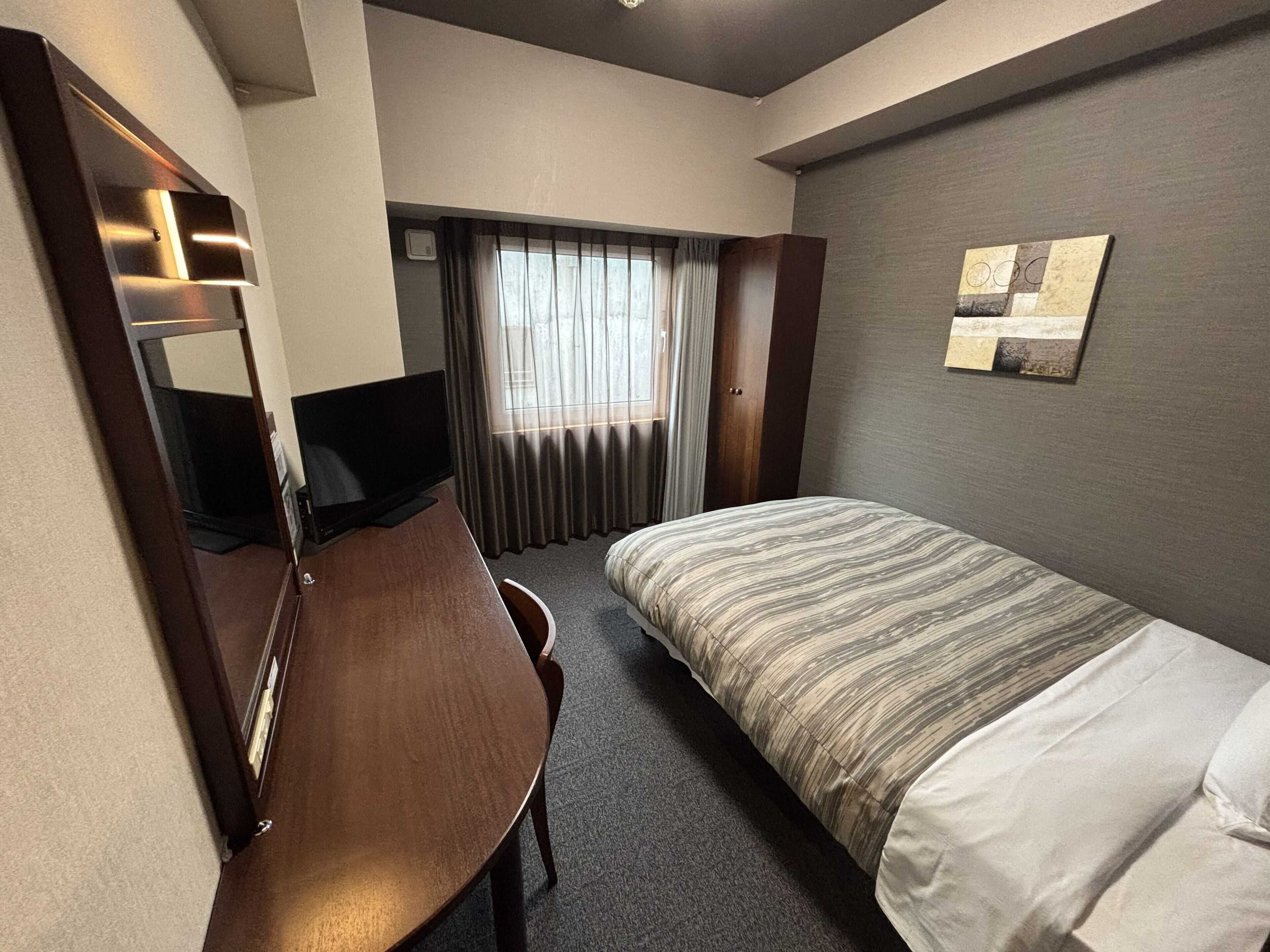 A hotel room with a bed, a desk, a television, a window with curtains, and a wall-mounted artwork.