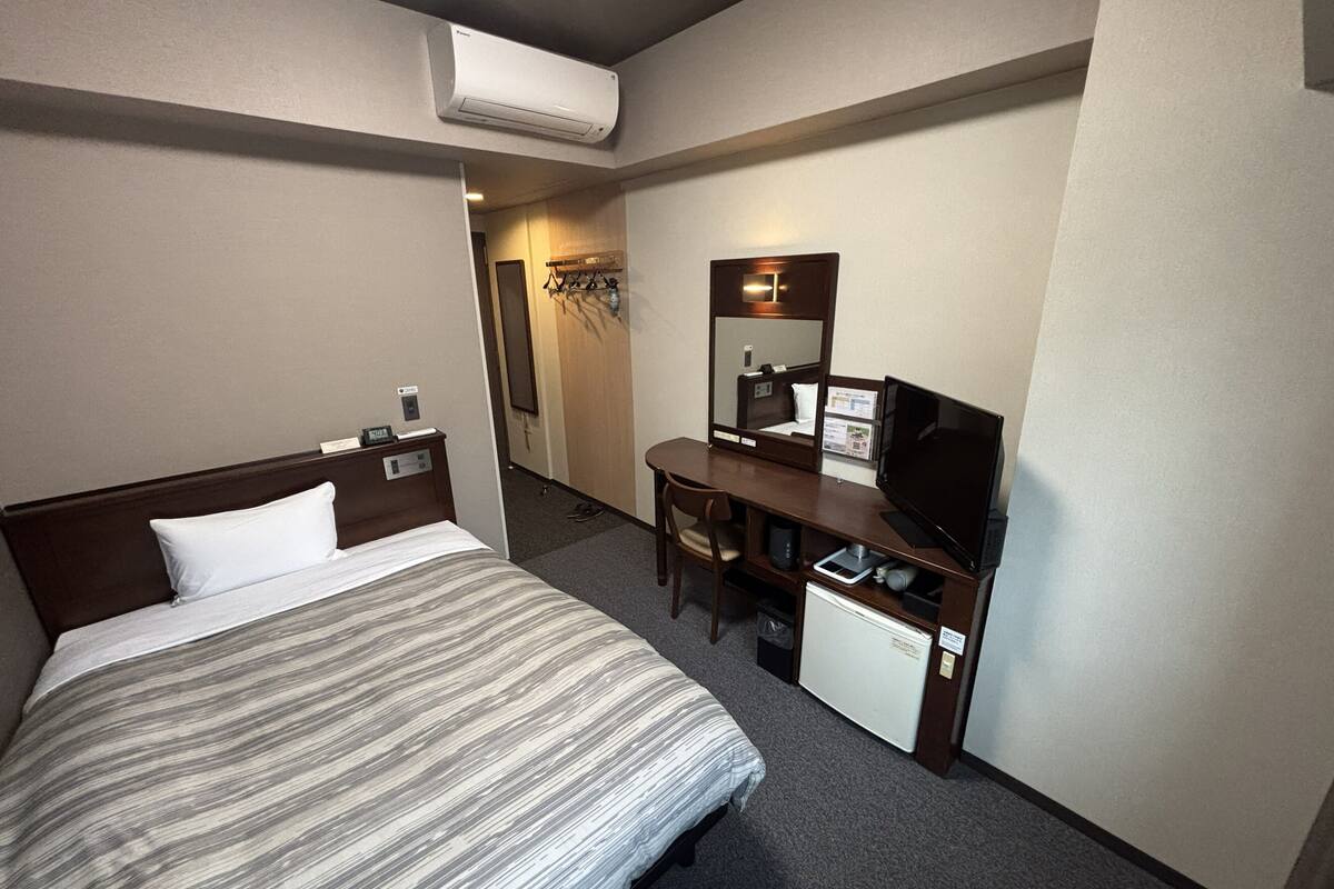 A hotel room with a bed, a desk with a television, a small refrigerator, and an air conditioning unit.