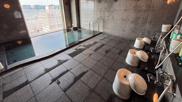 A modern bathroom with a glass-enclosed jacuzzi, a large window offering a city view, and a marble vanity with multiple sinks. - Hotel Route Inn Tokuyama Ekimae (Shunan)